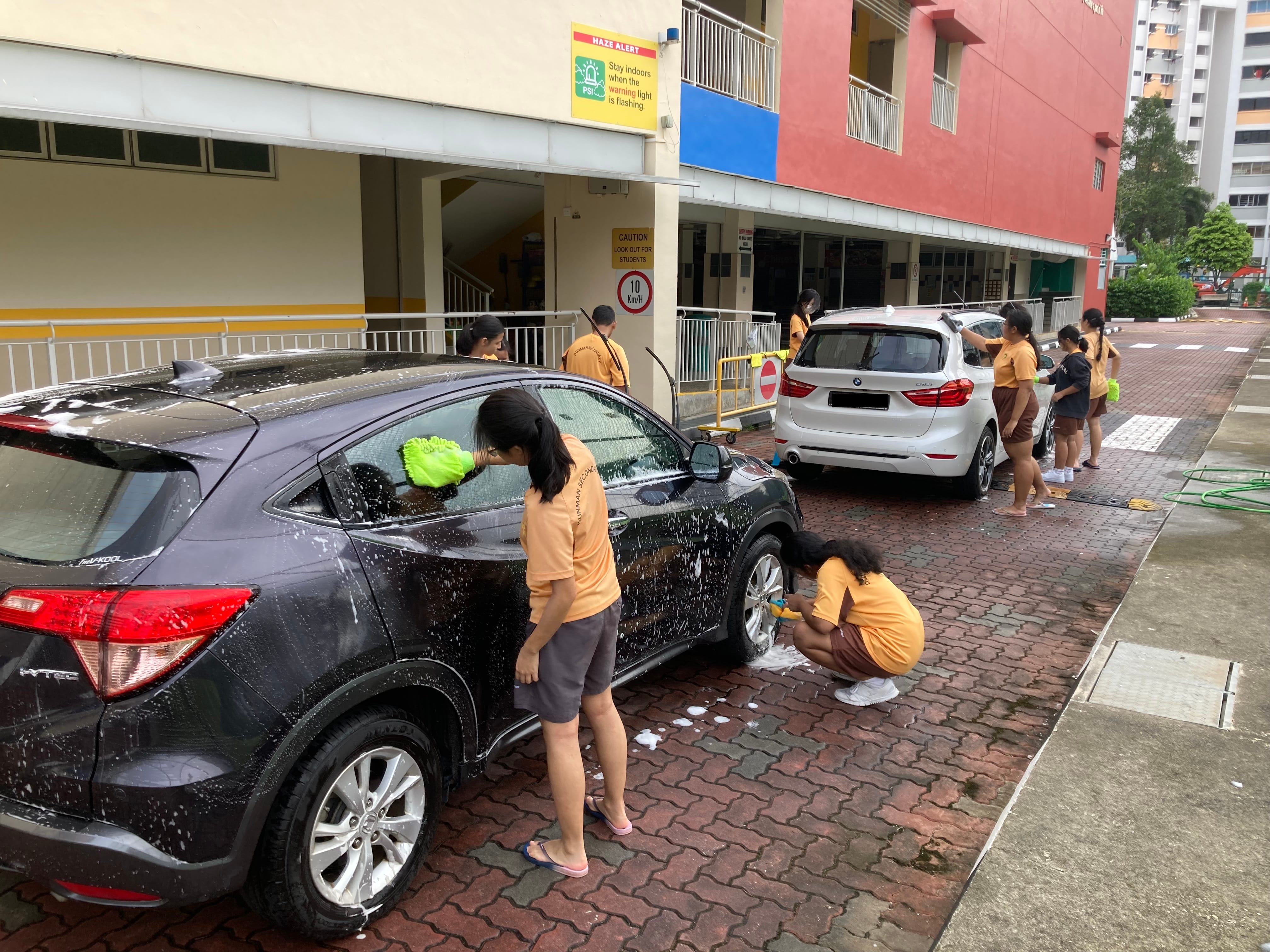 Students in orange uniforms scrubbing two soapy cars on a brick driveway outside a building.